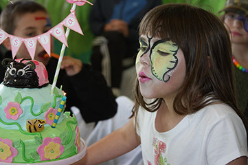 A young girls blows out candles on a beautifully decorated cake with flowers and bumble bees. Her face has a butterfly painted on it.