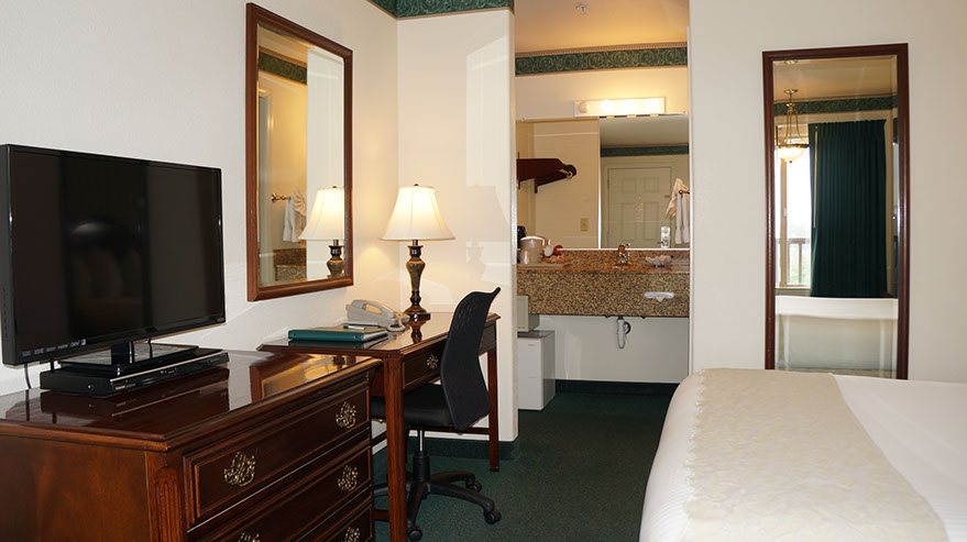 The Cozy Queen Guest Room Photo showing queen bed, dresser, flat screen tv, writing desk with lamp and direct dial phone. Entrance to dressing/wash area.