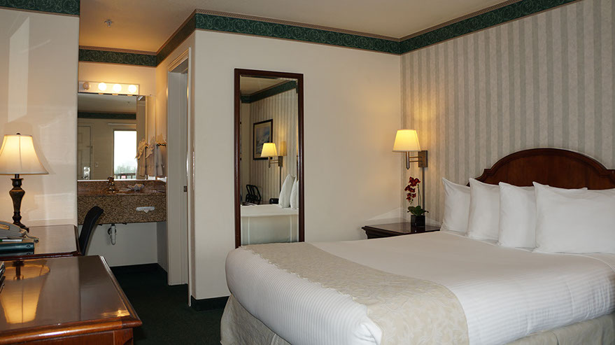 The Cozy Queen Guest Room Photo showing queen bed, dresser, writing desk with lamp, night stand with lamp, fulll length mirror, and entrance to dressing/wash area.