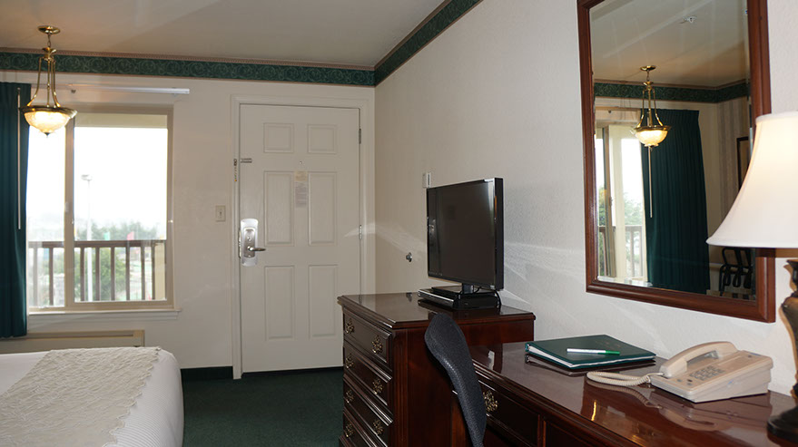 The Cozy Queen Guest Room Photo showing queen bed, dresser, flat screen tv, writing desk and chair, large mirror, direct dial phone, lamp and large window.