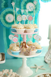 Beautifully decorated cupcakes rest upon a tier at a party. Decorated with soft pastels and seashells.